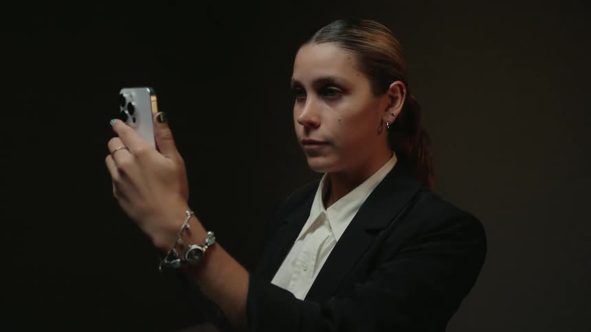 A focused woman in a business suit raises her smartphone in slow motion against a dark background