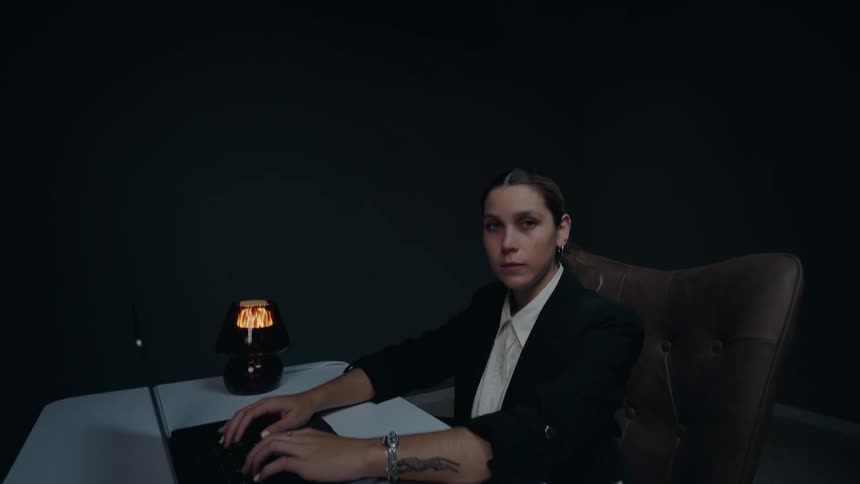 High-angle shot of a serious businesswoman working on her laptop in a dark, moody office, ending with her staring at the camera