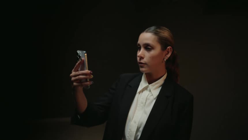 A stylish woman in a dark suit seriously gazes at her smartphone screen in a dimly lit room