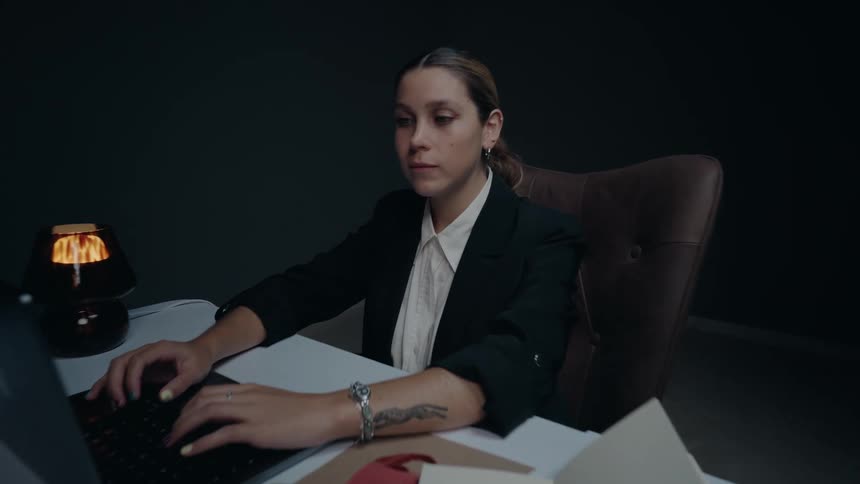 A businesswoman in a dark office works on her laptop, then stops and looks at the camera with a gentle smile