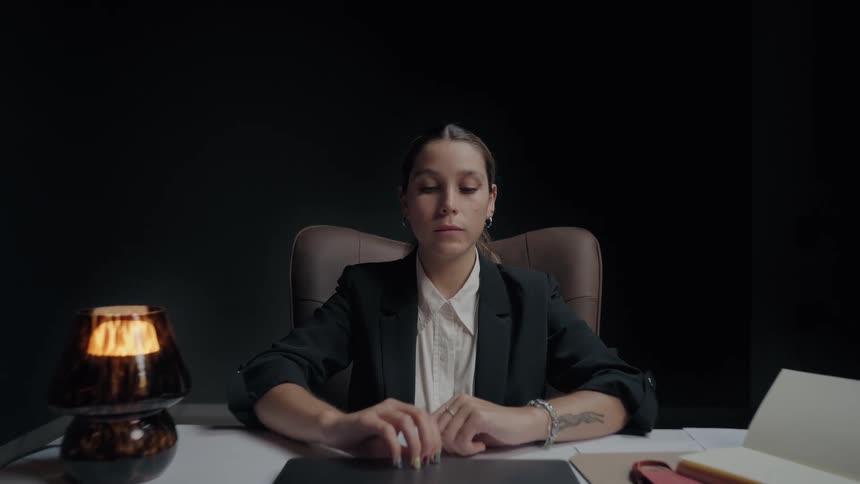 A serious businesswoman in a dark, modern office sits at her desk and slowly opens her laptop to begin working