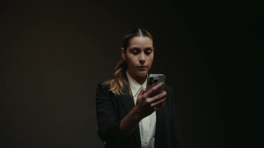 A serious businesswoman in a dark room picks up her smartphone and starts scrolling