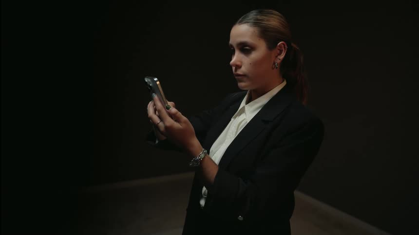 A young woman dressed in a business casual blazer scrolls through her smartphone in a dark, moody room