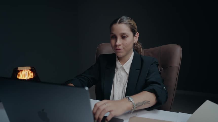 A businesswoman opens her laptop at her desk in a dark, modern office and begins to work with a smile