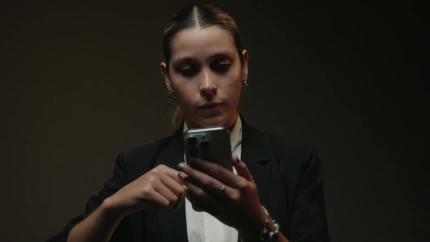 Stylish young woman dressed in a business suit scrolls through her phone in a dimly lit studio