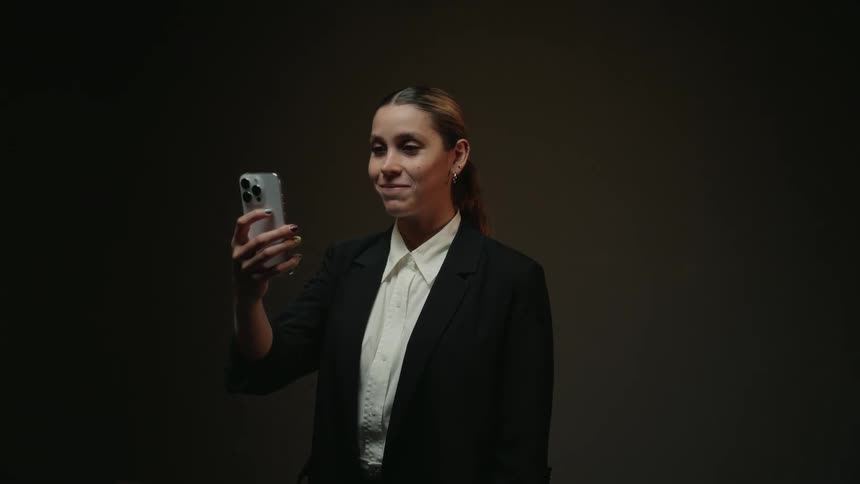 A woman in a black business suit smiles while looking at her smartphone in slow motion against a dark background