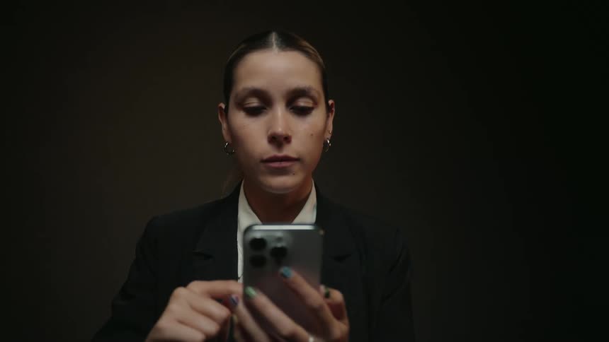 A focused businesswoman in a black suit uses her smartphone in a dark studio as the camera slowly pushes in