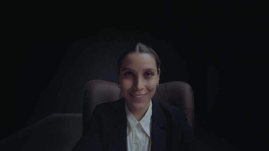 A businesswoman works on her laptop in a dark room, then looks up and smiles directly at the camera as it zooms in
