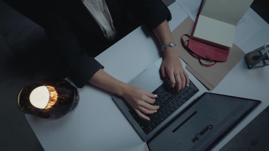A top-down shot zooms out from a businesswoman's hands typing on a laptop to reveal her working late at her desk by candlelight