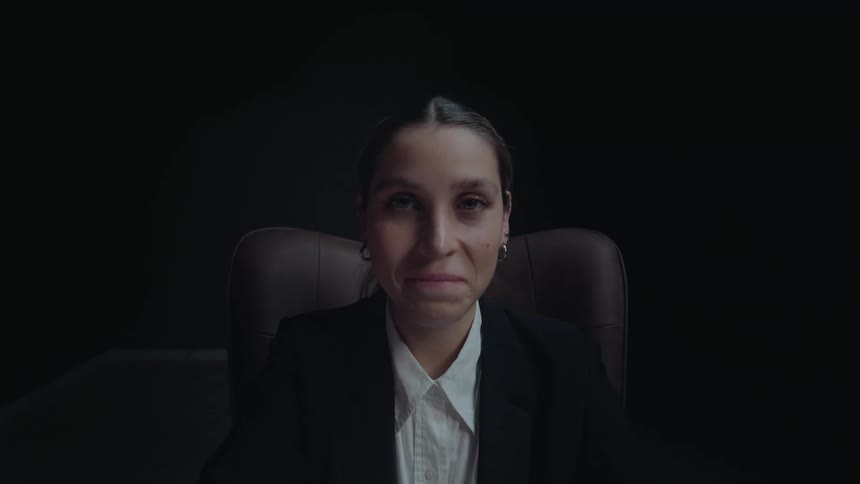 A businesswoman works on a laptop in a dark office, then looks up from her desk and smiles warmly at the camera