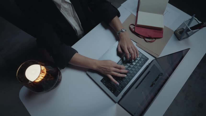 Overhead shot zooms out from a businesswoman's hands typing on a laptop as she works late at night
