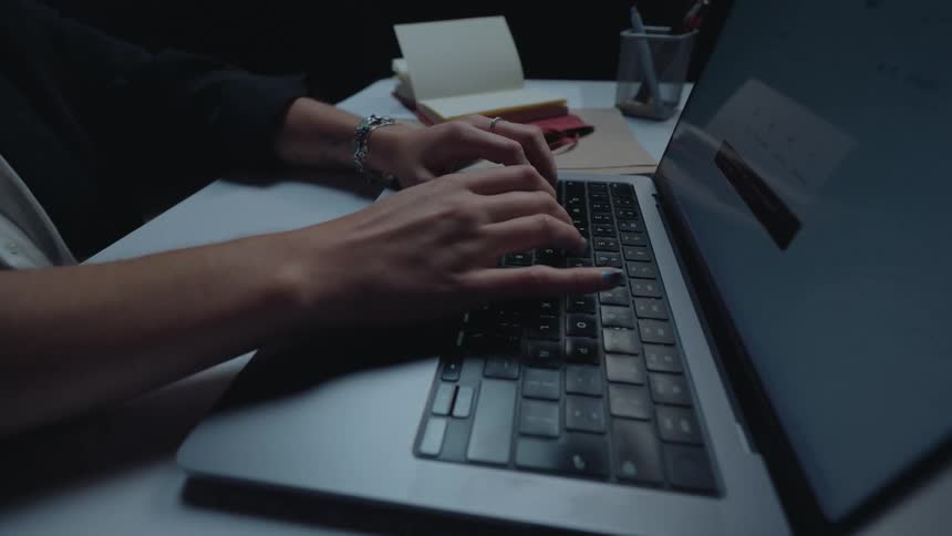 A close-up shot of a woman's hands typing on a laptop keyboard late at night in a dark office
