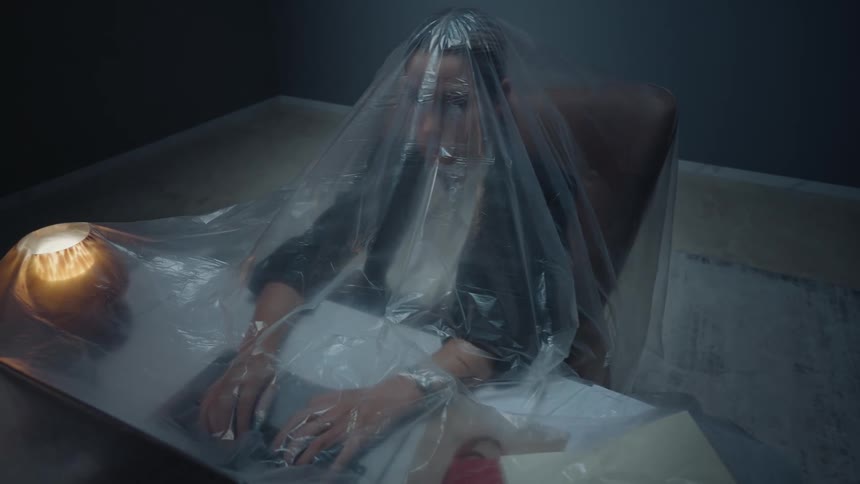 A woman covered by a large plastic sheet stops working on her laptop in a dark, candlelit room, symbolizing isolation and claustrophobia