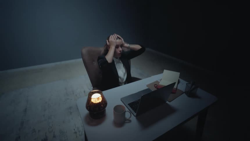 An overhead dolly-out shot reveals a frustrated businesswoman holding her head while working late at her desk in a dark room