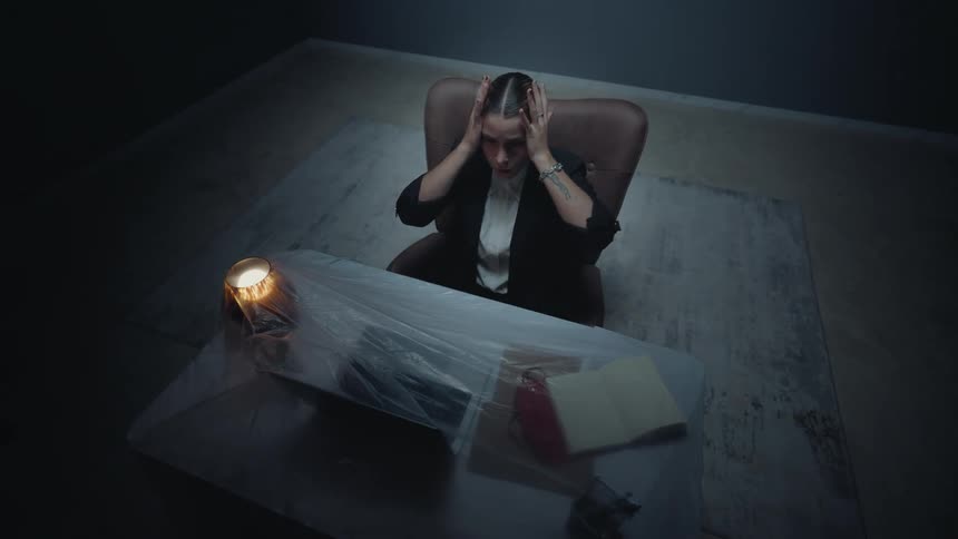 A high-angle view shows an anxious businesswoman in a dark, surreal office, holding her head in distress at a desk covered in plastic