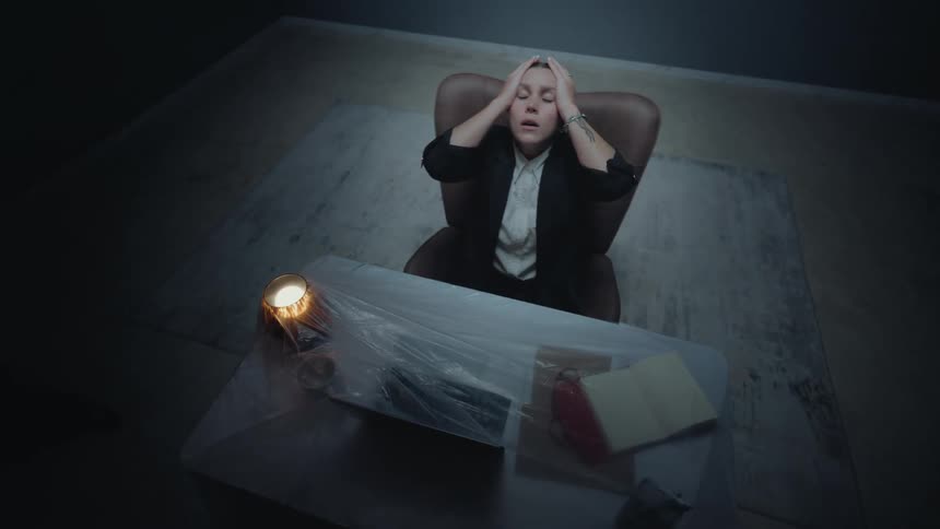 A stressed woman sits at her desk in a dark room, overcome with anxiety and holding her head in her hands