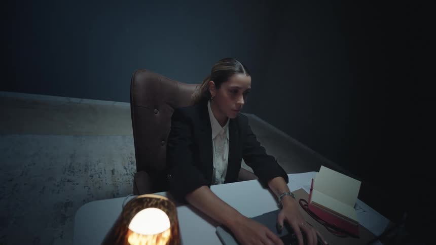 A determined businesswoman stays late in a dark, isolated office, diligently working on her laptop under the warm glow of a desk lamp