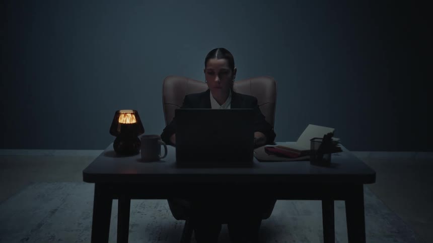 A focused businesswoman works late at her desk in a dark, minimalist office, illuminated only by a small lamp and her laptop