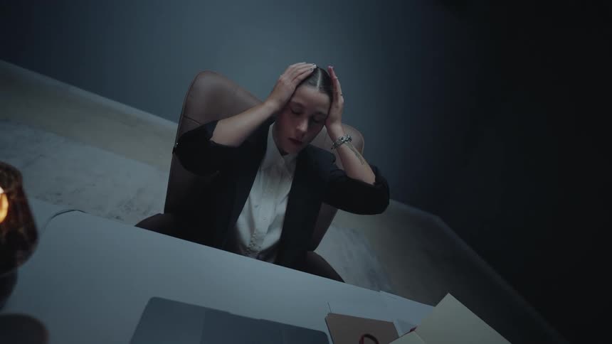 A stressed businesswoman working late at night in a dimly lit office feels overwhelmed and clutches her head