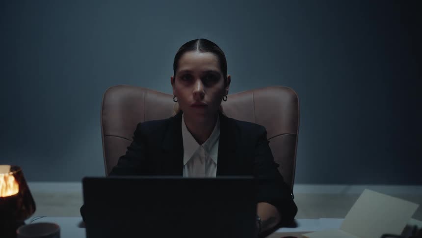 A focused businesswoman works late at her desk in a dark, moody office, then stops and stares intently at the camera