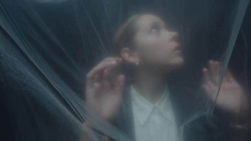 A distressed young woman trapped behind a translucent plastic sheet presses her hands against it in slow motion