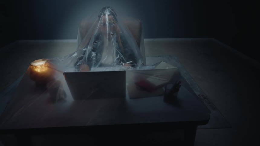 An unsettling shot of a person working on a laptop in a dark room, completely enveloped with their desk under a large plastic sheet
