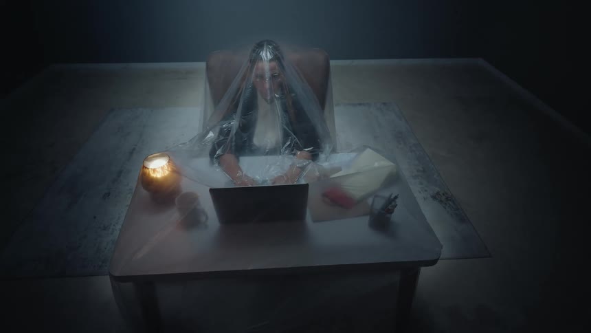 Surreal high-angle shot of a woman working on a laptop in a dark office, trapped and isolated under a large plastic sheet