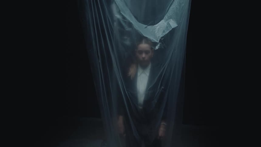 A still portrait of a young woman in a business suit standing behind a sheer plastic curtain in a dark, moody room