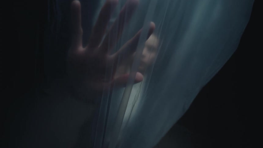 A frightened woman trapped behind a translucent plastic sheet presses her hand against it in a dark, atmospheric setting