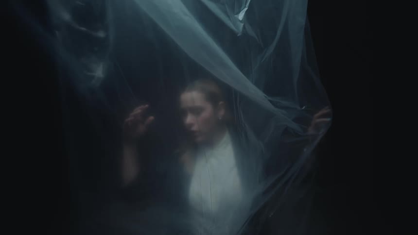 A woman is slowly enveloped by a sheet of plastic in a dark room, conveying a sense of anxiety and being trapped