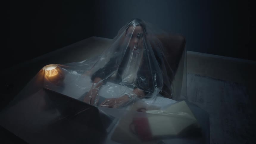 A surreal high-angle dolly shot shows a woman working on her laptop at a desk, trapped under a large plastic sheet in a dark, empty room