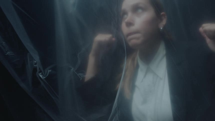 A young woman in business attire appears trapped and anxious behind a sheer plastic curtain in a dark room