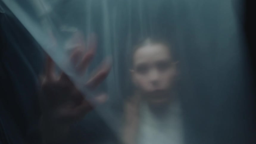 A mysterious woman moves gracefully behind a blurry, translucent plastic sheet in a dark, dreamlike setting