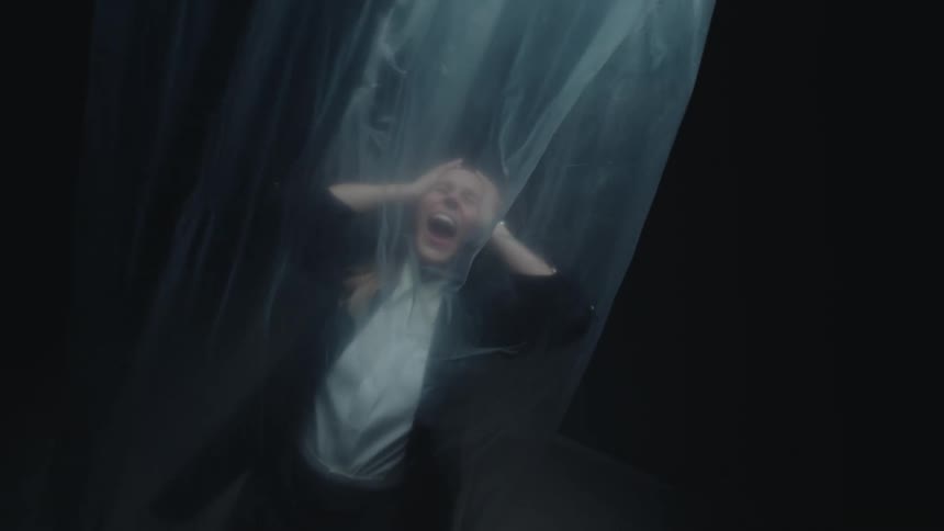 A terrified woman trapped behind a sheet of plastic in a dark room grabs her head and screams in agony