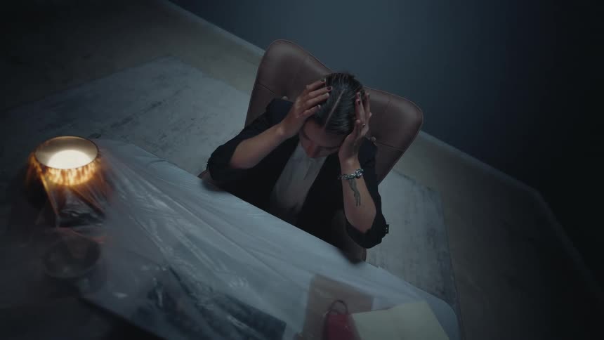 A stressed businesswoman holds her head in despair while sitting at a plastic-covered desk in a dark, abandoned office