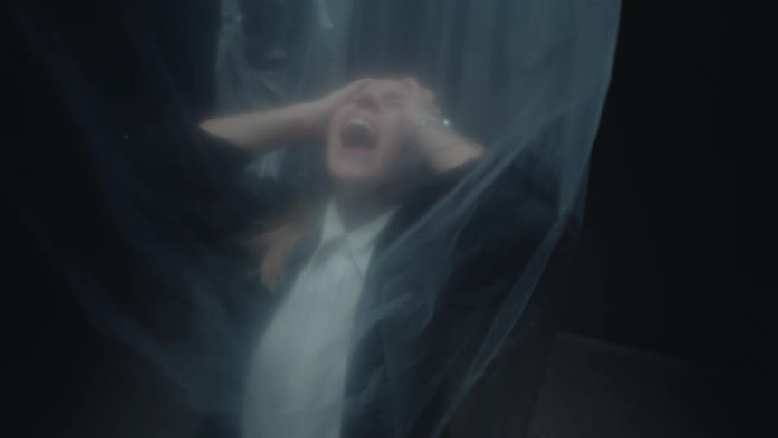 A terrified person in a suit screams in agony and despair while trapped behind a translucent plastic sheet in a dark room