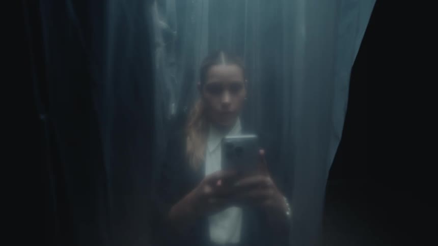 Moody close-up shot of a young woman taking a selfie on her smartphone, viewed through a blurry plastic sheet in a dark, mysterious setting