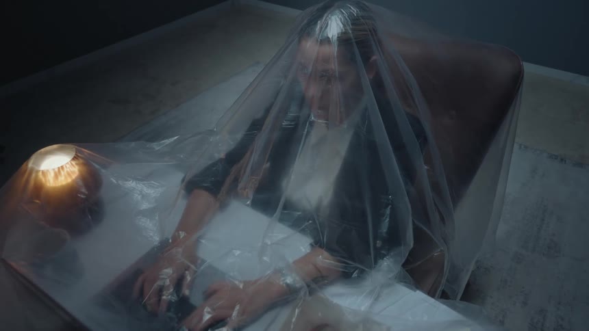 A surreal high-angle shot captures a businesswoman trapped under a plastic sheet, working on her laptop in a dark room