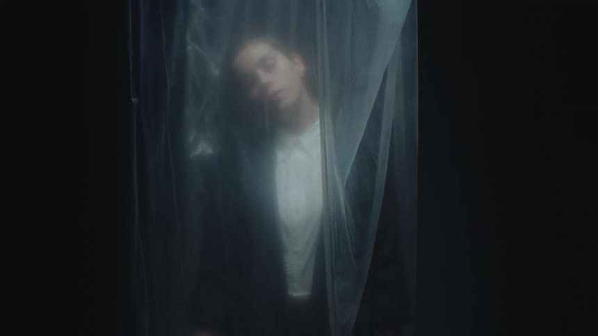 A mysterious woman in a business suit is seen behind a translucent plastic sheet in a dark, atmospheric setting