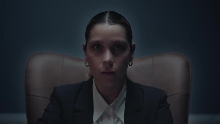 A serious businesswoman with an intense gaze sits in a dark room, staring directly at the camera