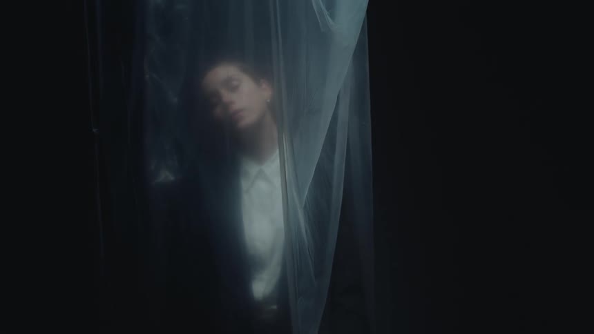 A somber woman in a suit slowly lowers her head behind a translucent plastic sheet in a dark, atmospheric setting