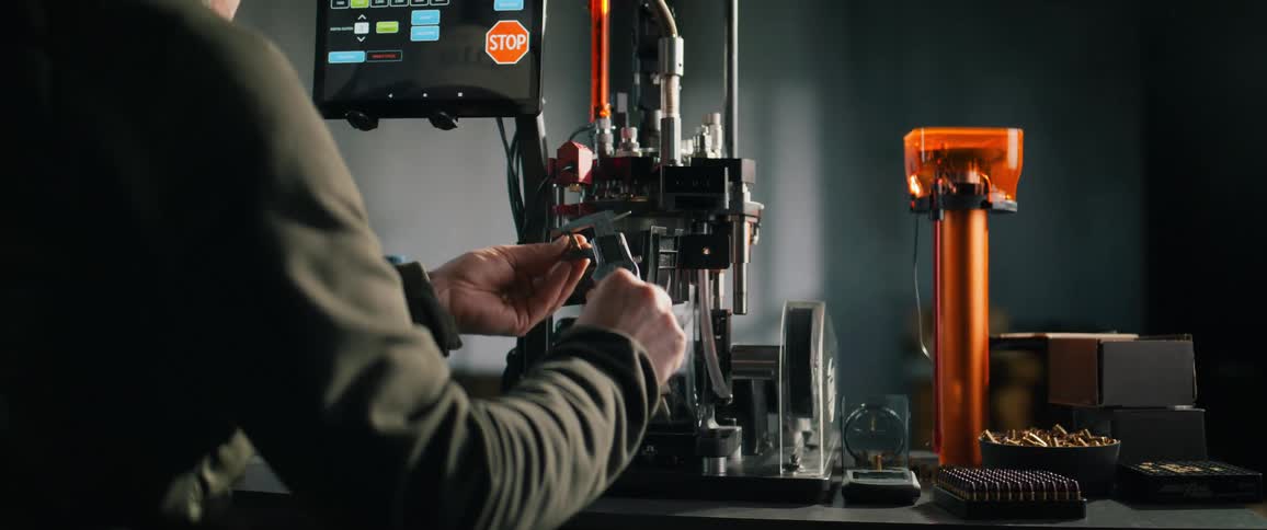 A man operates an automated ammunition reloading press in his workshop, carefully measuring a finished cartridge with a caliper for quality control