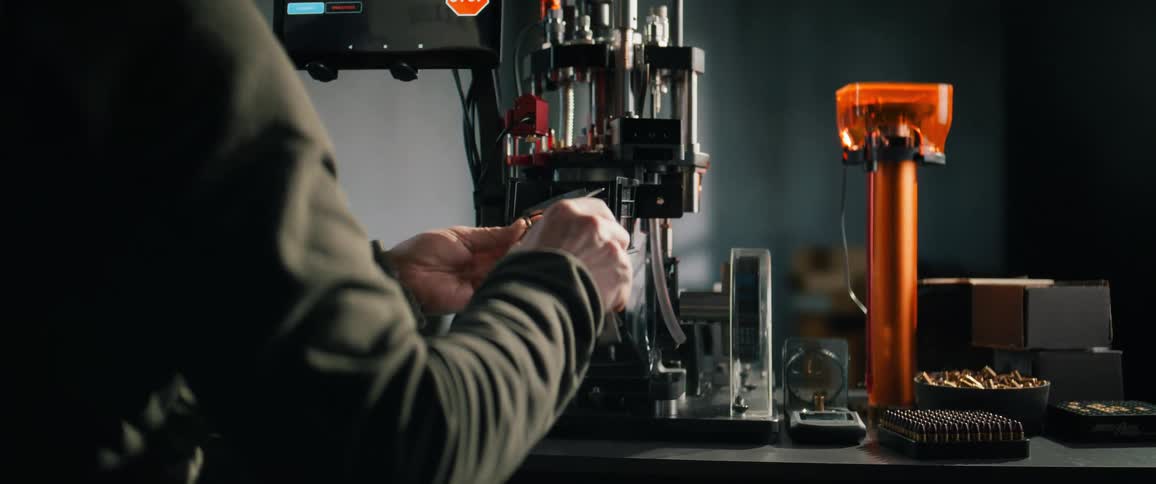 A skilled craftsman operates an automated ammunition reloading press, carefully measuring a bullet casing with calipers in his workshop