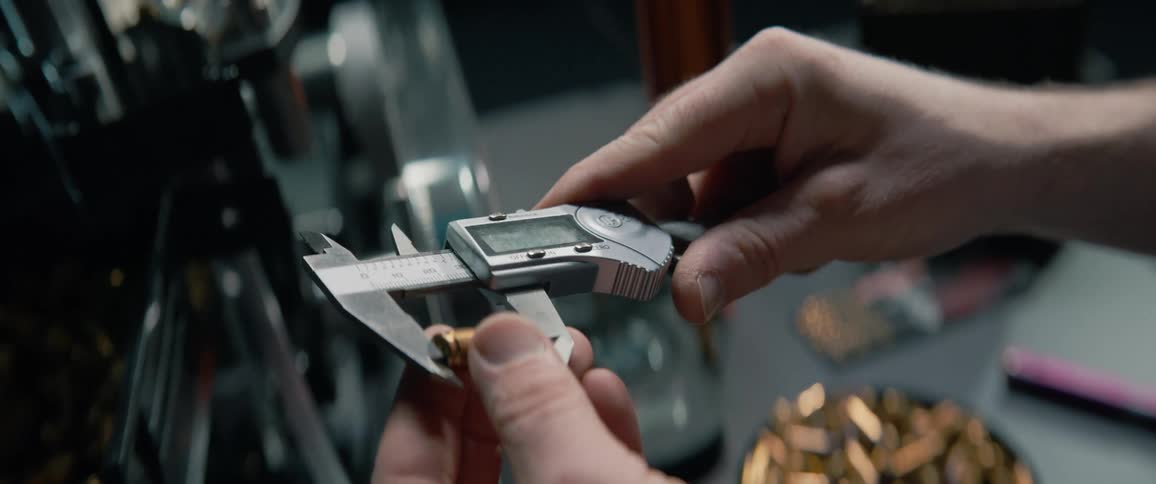 A close-up shot shows a man meticulously measuring a bullet casing with a digital caliper as part of the ammunition reloading process