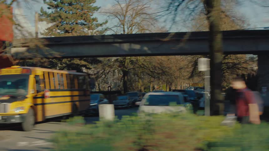 A dreamy, motion-blurred tracking shot past pedestrians and a school bus on a sunlit city street