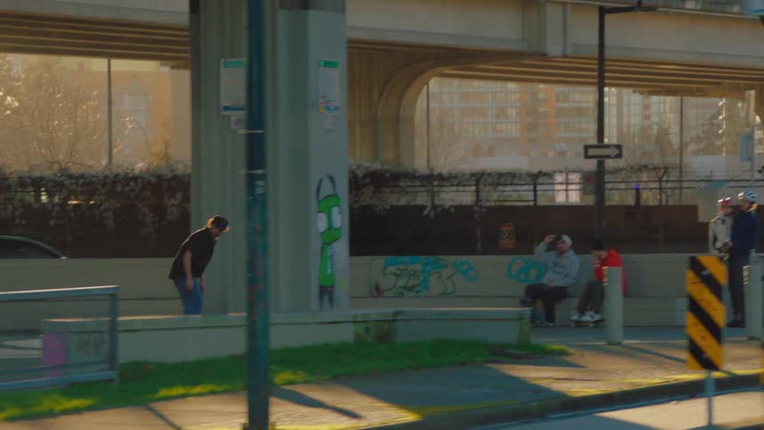 A skateboarder in slow motion grinds a rail at an urban skatepark under an overpass during golden hour