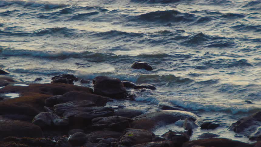 Moody slow motion footage shows ocean waves crashing over dark rocks on the shore