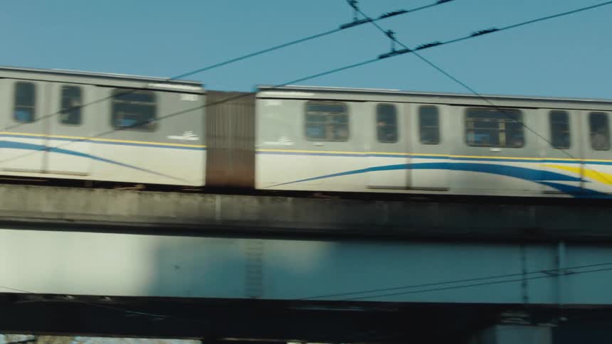 A side-on tracking shot follows a Vancouver SkyTrain as it travels along an elevated track on a clear sunny day