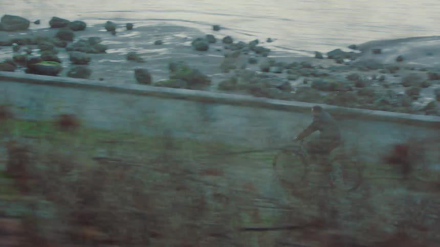 Parallax tracking shot reveals a man cycling on a path along a rocky coastline
