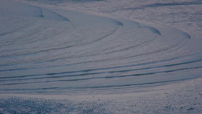 A telephoto shot captures the wind whipping snow across a vast and frozen arctic landscape, creating textured drifts